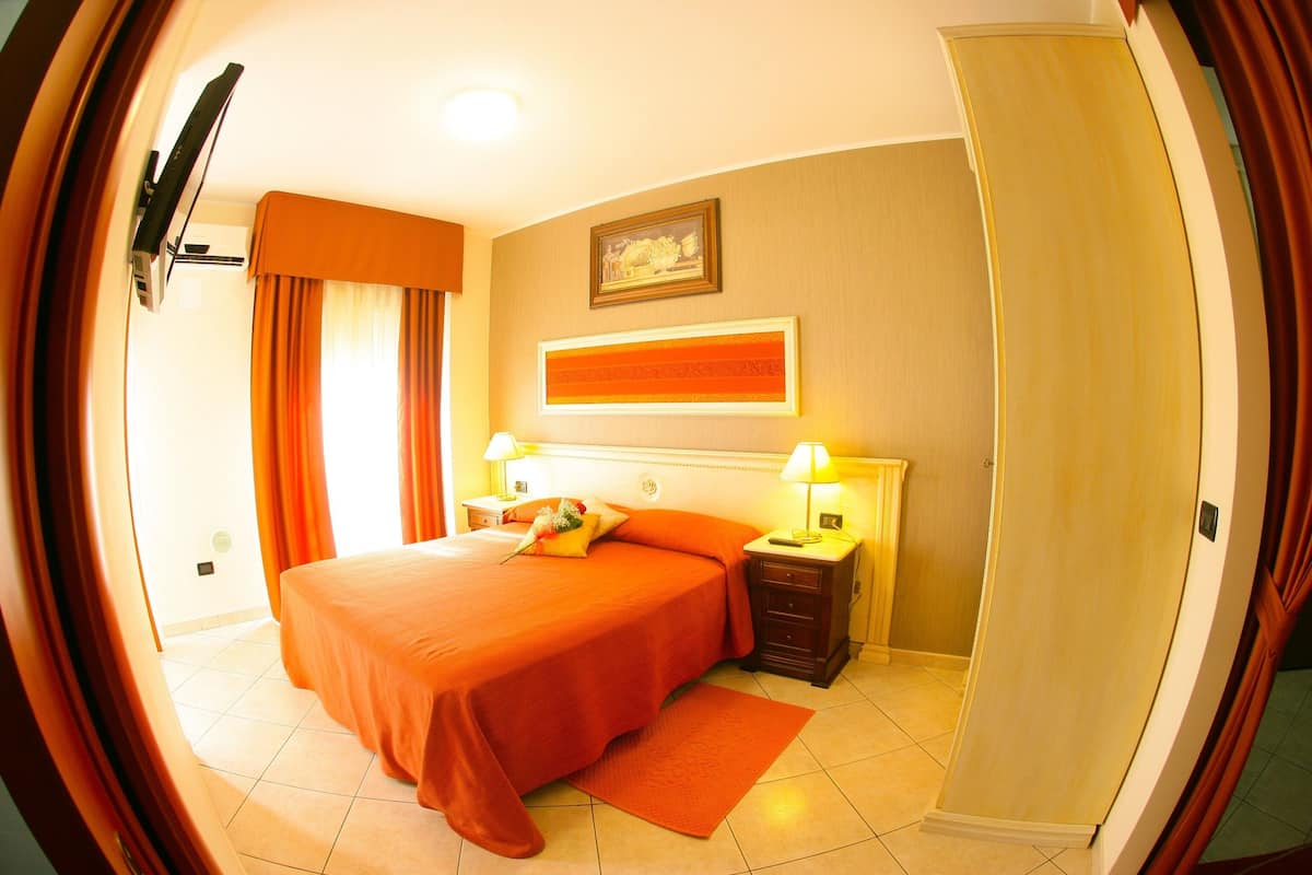 A hotel room with a bed, two bedside tables with lamps, a window with curtains, a painting on the wall, and a door.