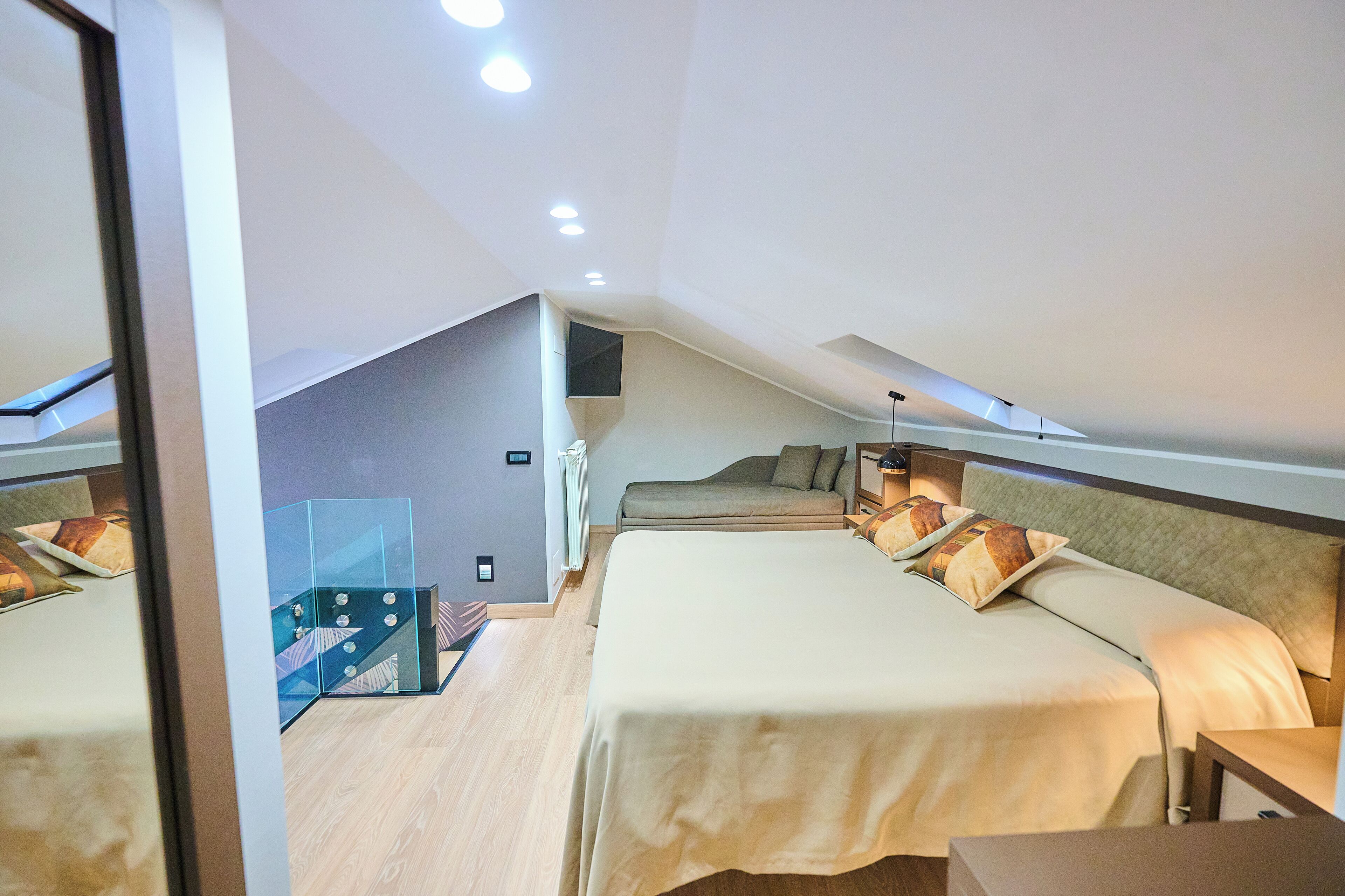 A modern hotel room with a sloped ceiling, two beds, a seating area, and a glass partition.