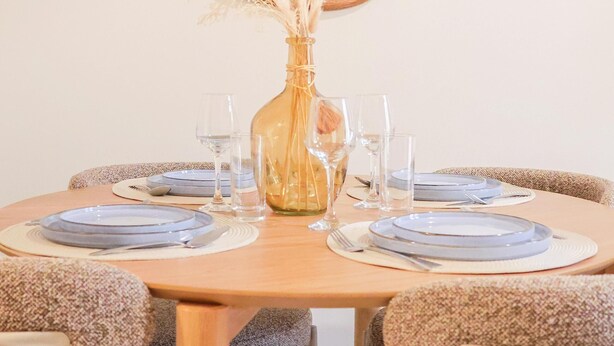Ein runder Esstisch mit Holzrahmen, umgeben von vier gepolsterten Stühlen. Auf dem Tisch steht eine Vase mit Pampasgras.