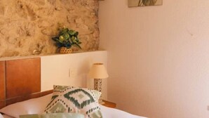 A room with a stone wall, a bed with patterned pillows, a small plant, and a wall-mounted lamp. - Cosy gîte for 6 people in the heart of the Fontfroide massif (Narbonne)