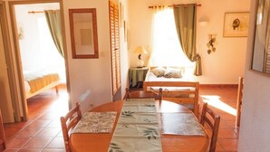 A dining area with a round table, chairs, and a tablecloth. There is a bed with a window in the background. - Cosy gîte for 6 people in the heart of the Fontfroide massif (Narbonne)