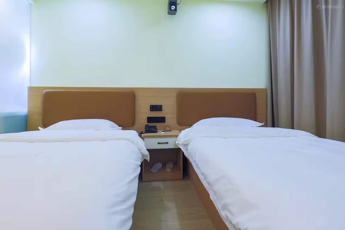 A hotel room with two single beds, a wooden headboard, a nightstand with a phone and a small clock, and a window with curtains.