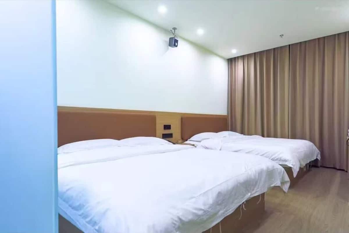 A hotel room with two single beds, a wooden headboard, a ceiling-mounted projector, and a large window with curtains.