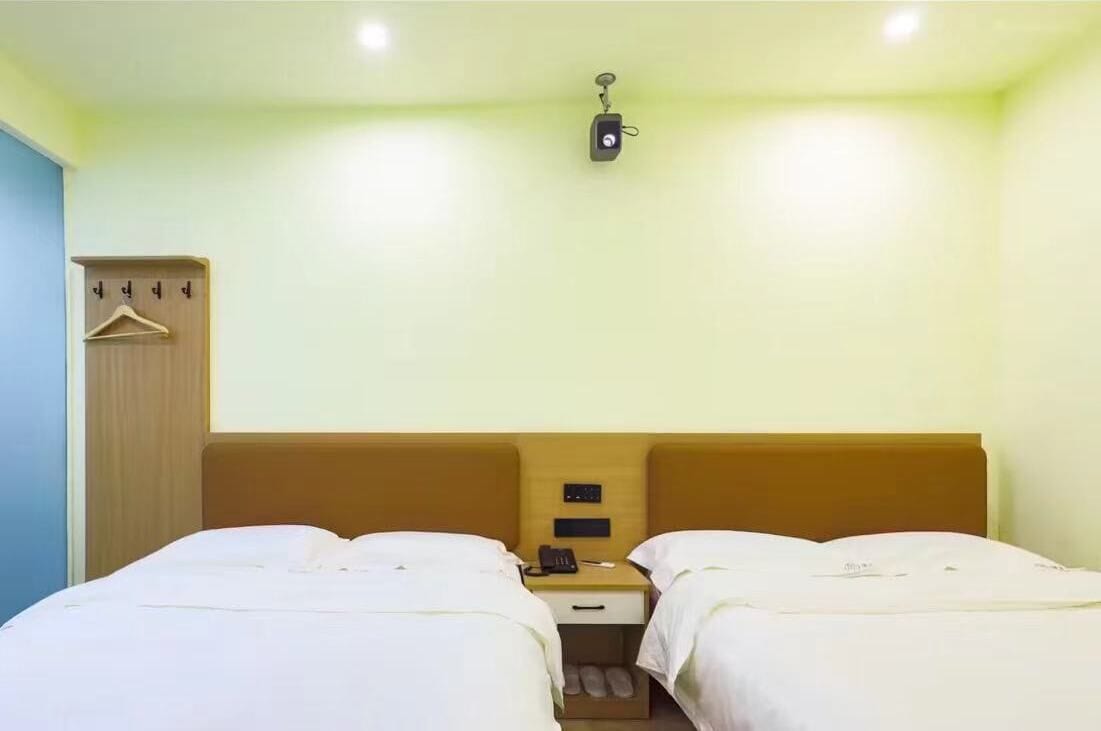 A hotel room with two beds, a wooden wardrobe, a small desk with a phone, and a wall-mounted light fixture.