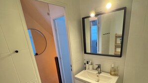 Un bagno con lavandino, specchio e servizi igienici bianchi. Si apre una porta verso un'altra stanza.