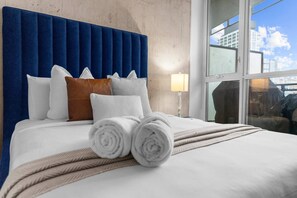 Ett hotellrum med en stor säng, en blå klädd sänggavel och utsikt över staden genom fönstret.