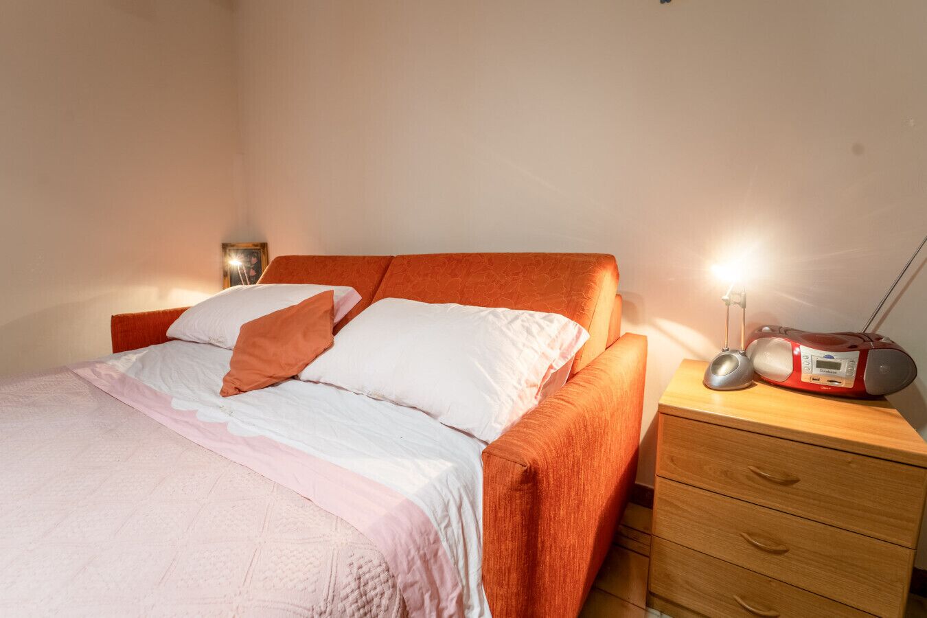 Een bed met een oranje hoofdbord, een nachtkastje met een radio en een lamp.