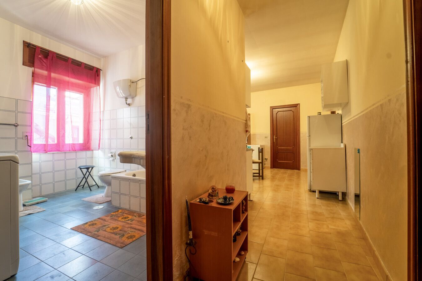 A narrow hallway with a bathroom on the left, featuring a toilet, sink, and bathtub. A wooden cabinet with various items is visible in the hallway.