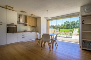 Eine moderne Küche mit weißen Schränken, ein Essbereich mit Tisch und Stühlen und eine große Glasschiebetür, die auf eine Terrasse mit Blick auf Grünfläche führt.