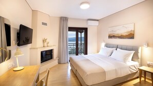 A modern hotel room with a large bed, a flat-screen TV, a desk with a lamp, a fireplace, and a balcony with a view. - Lake Kremasta Hotel (Agrafa)