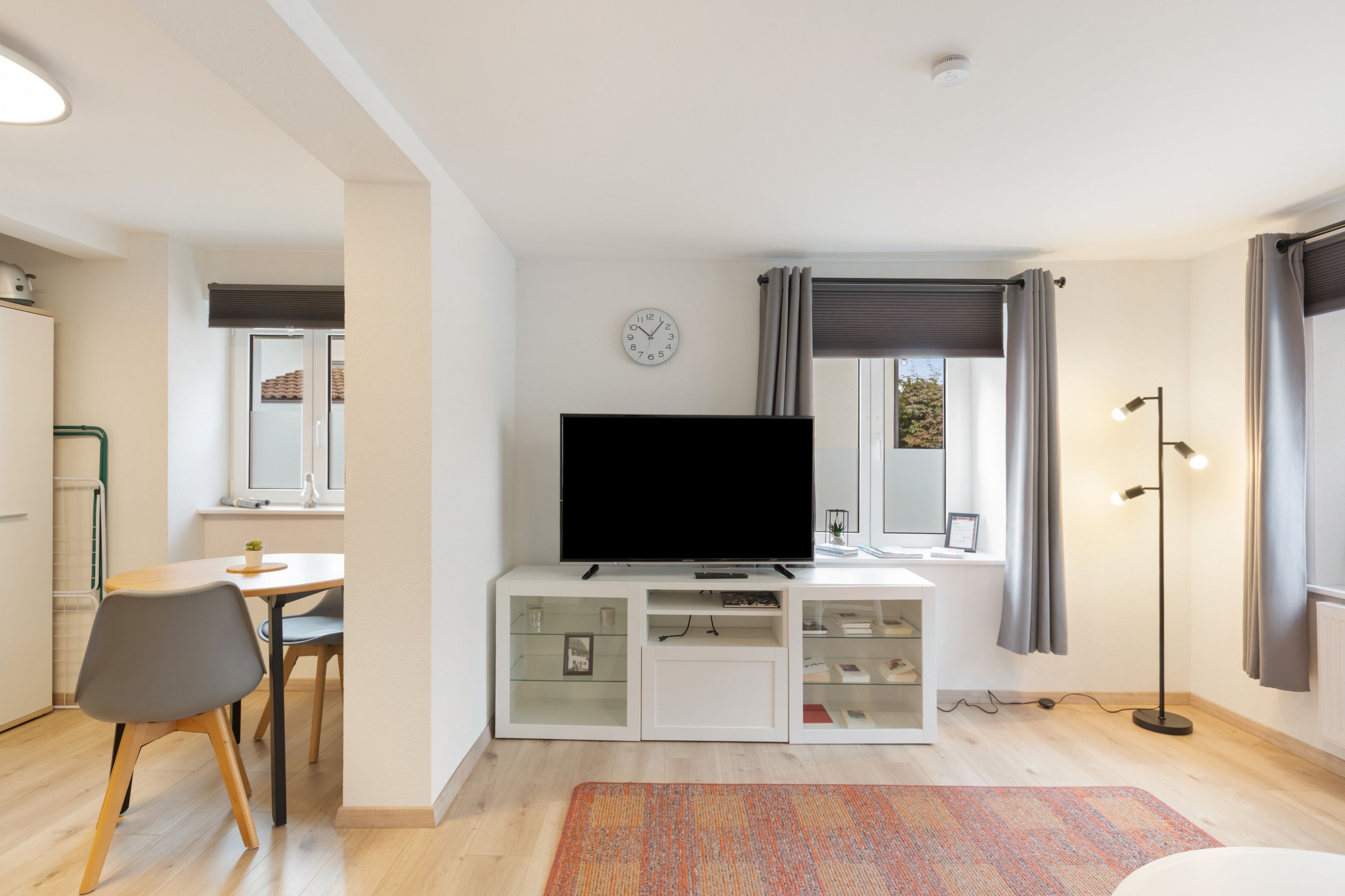 Una sala moderna con un televisor de pantalla plana sobre un mueble blanco, un comedor con mesa y sillas, y un ventanal con cortinas.
