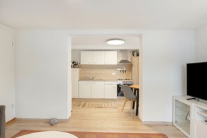 Cocina moderna con armarios blancos, estufa y lavamanos. Hay un comedor con mesa y sillas, y un televisor sobre un mueble.