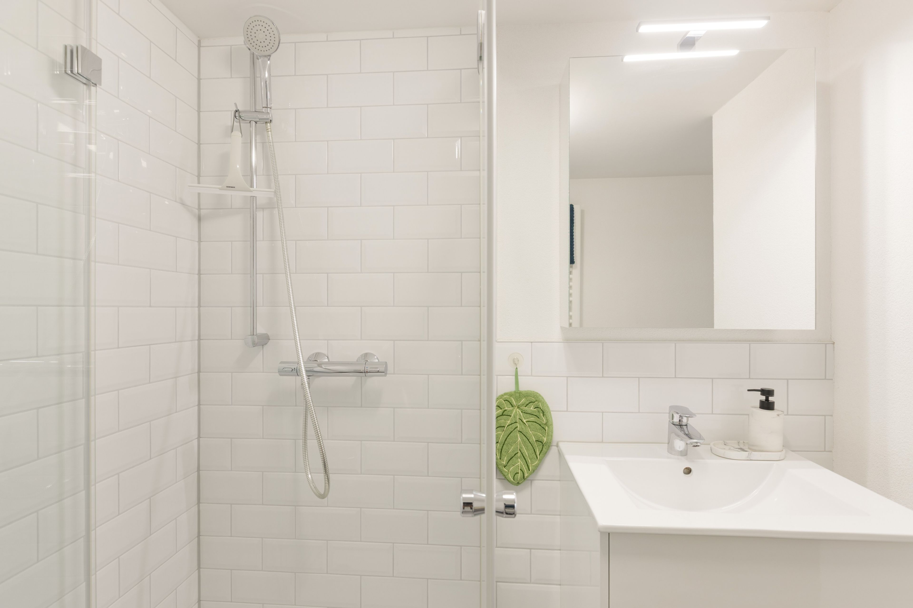 Ein modernes Badezimmer mit einer in Glas abgeschlossenen Dusche, weiß gefliesten Wänden, einem Waschbecken mit Spiegel und einem handtuchhalter in Form eines grünen Blattes.