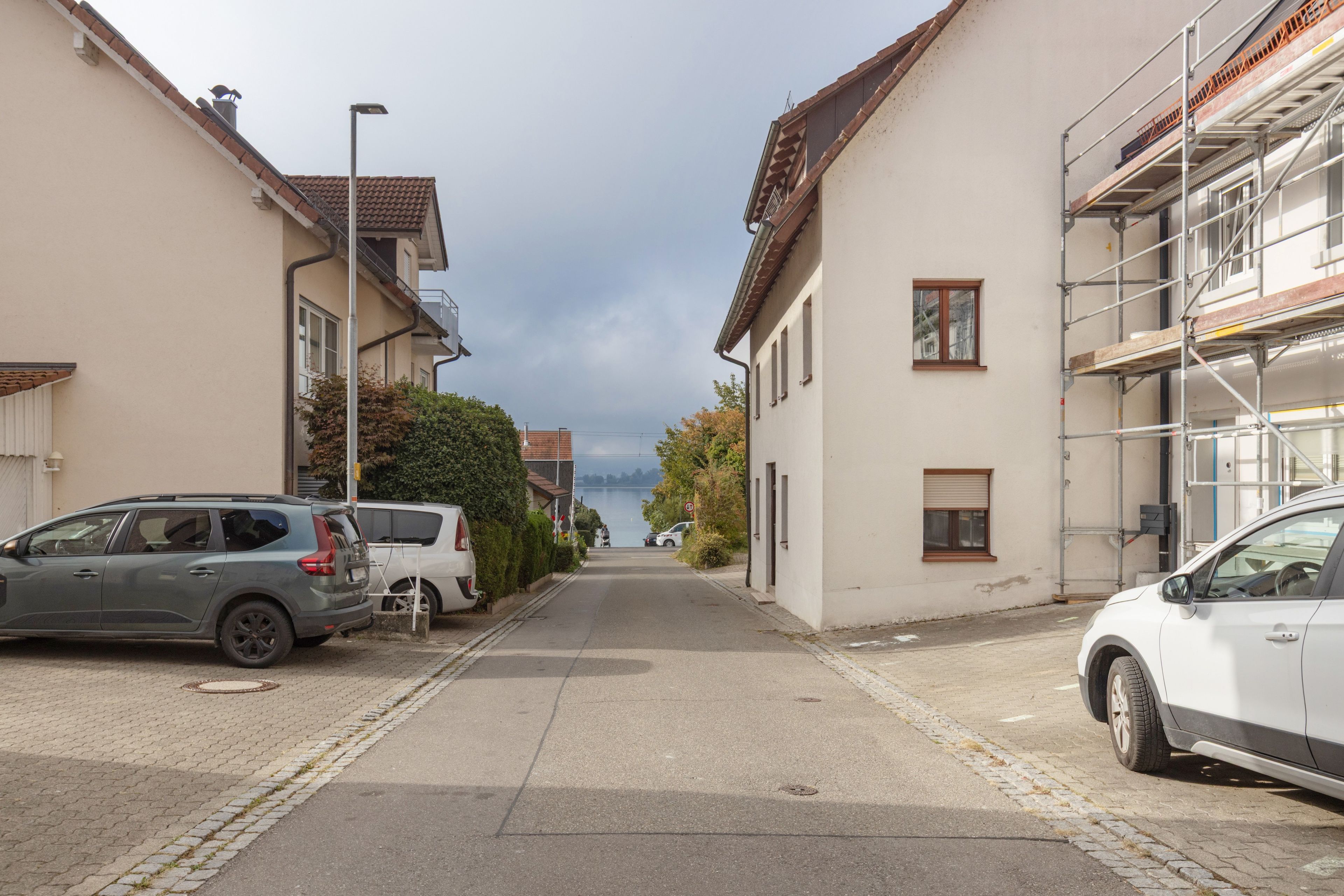 Una calle residencial con coches aparcados, casas y una vista despejada de un lago en la distancia.