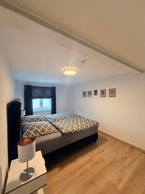 Ein Schlafzimmer mit Bett, Nachttisch mit Lampe, Fenster mit Vorhängen und Deckenleuchte.