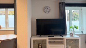 A modern living room with a flat-screen TV on a white entertainment unit, a dining area with a table and chairs, and a window with a view of greenery. - Apartment "Fewo Im Schönen Allensbach" with Lake View and Wi-Fi (Allensbach)