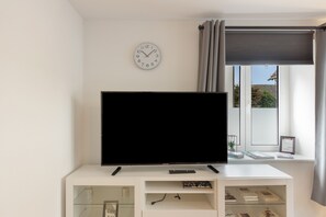 A living room with a flat-screen TV on a white stand, a clock on the wall, and a window with a view of the outside. - Apartment "Fewo Im Schönen Allensbach" with Lake View and Wi-Fi (Allensbach)