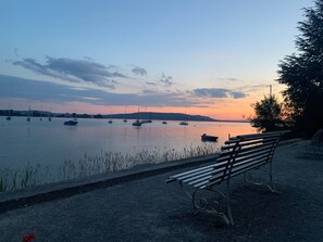Un banco junto a un cuerpo de agua tranquilo con barcos, cielo despejado y colinas al fondo.