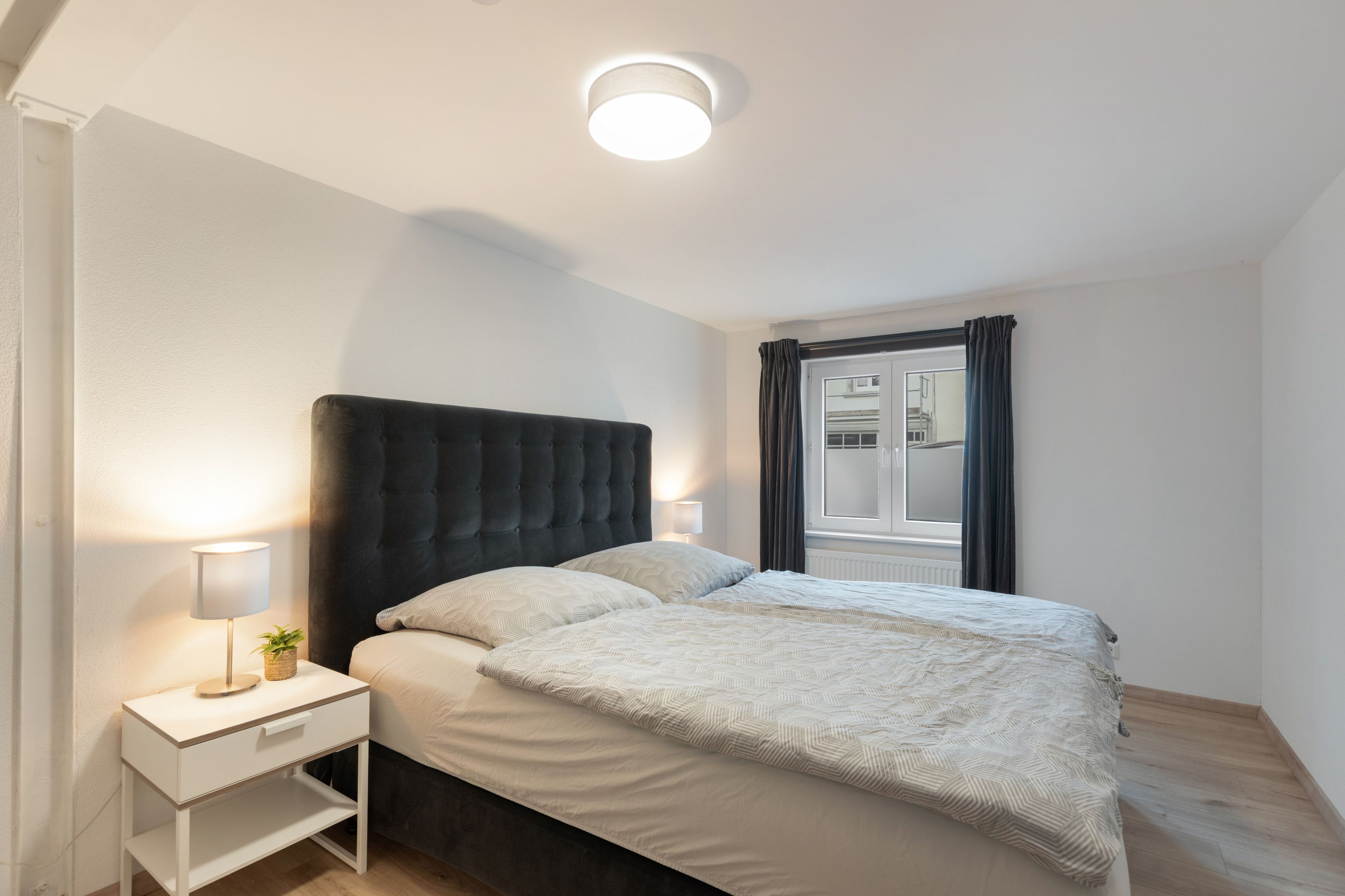 A bedroom with a bed, bedside table, lamp, and a window with curtains.