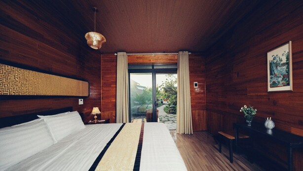 A bedroom with a large bed, wooden walls, a window with curtains, and a small table with a vase. - Havenora Retreat (Tay Hoa Lu)