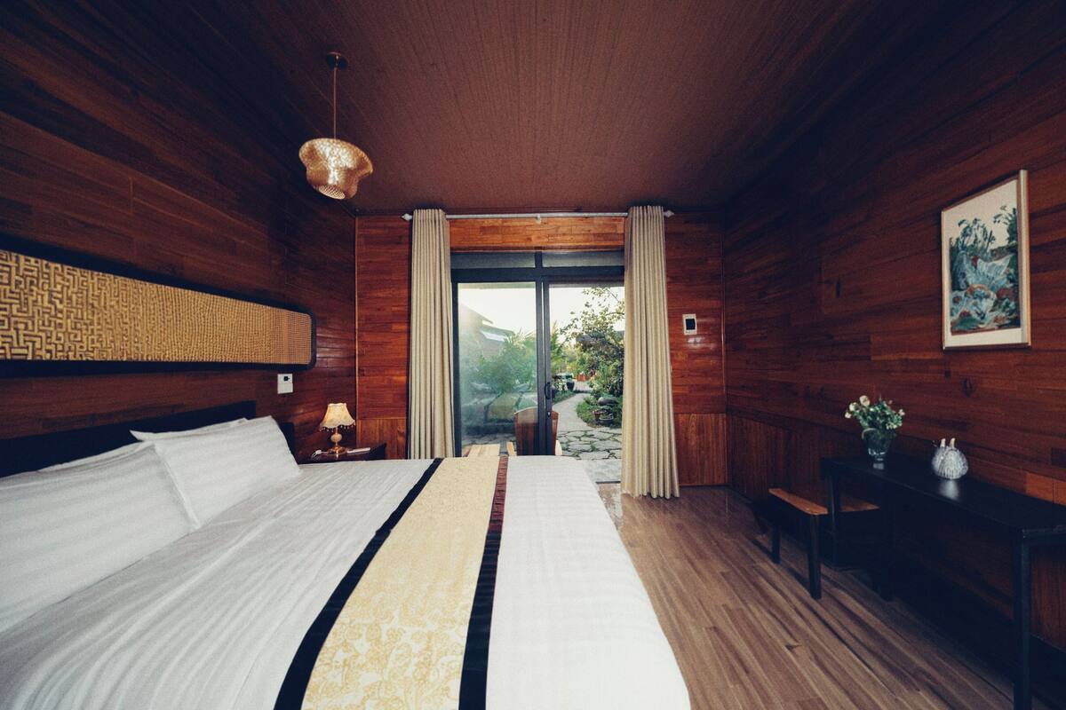 A bedroom with a large bed, wooden walls, a window with curtains, and a small table with a vase.