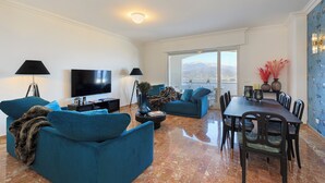 A modern living room with a large flat-screen TV, two sofas, a dining table with chairs, and a large window offering a mountain view. - Lake 270 Apartment (stresa)