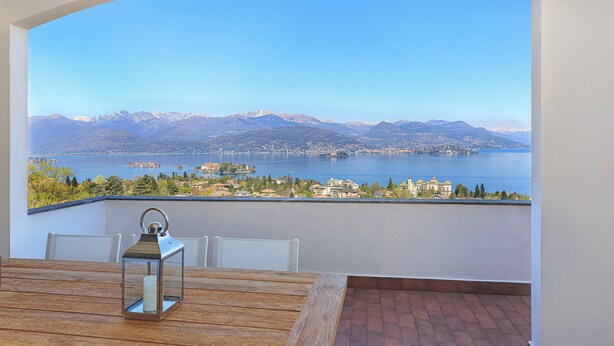 A balcony with a wooden table and chairs, a metal lantern, and a scenic view of mountains and a lake. - Lake 270 Apartment (stresa)