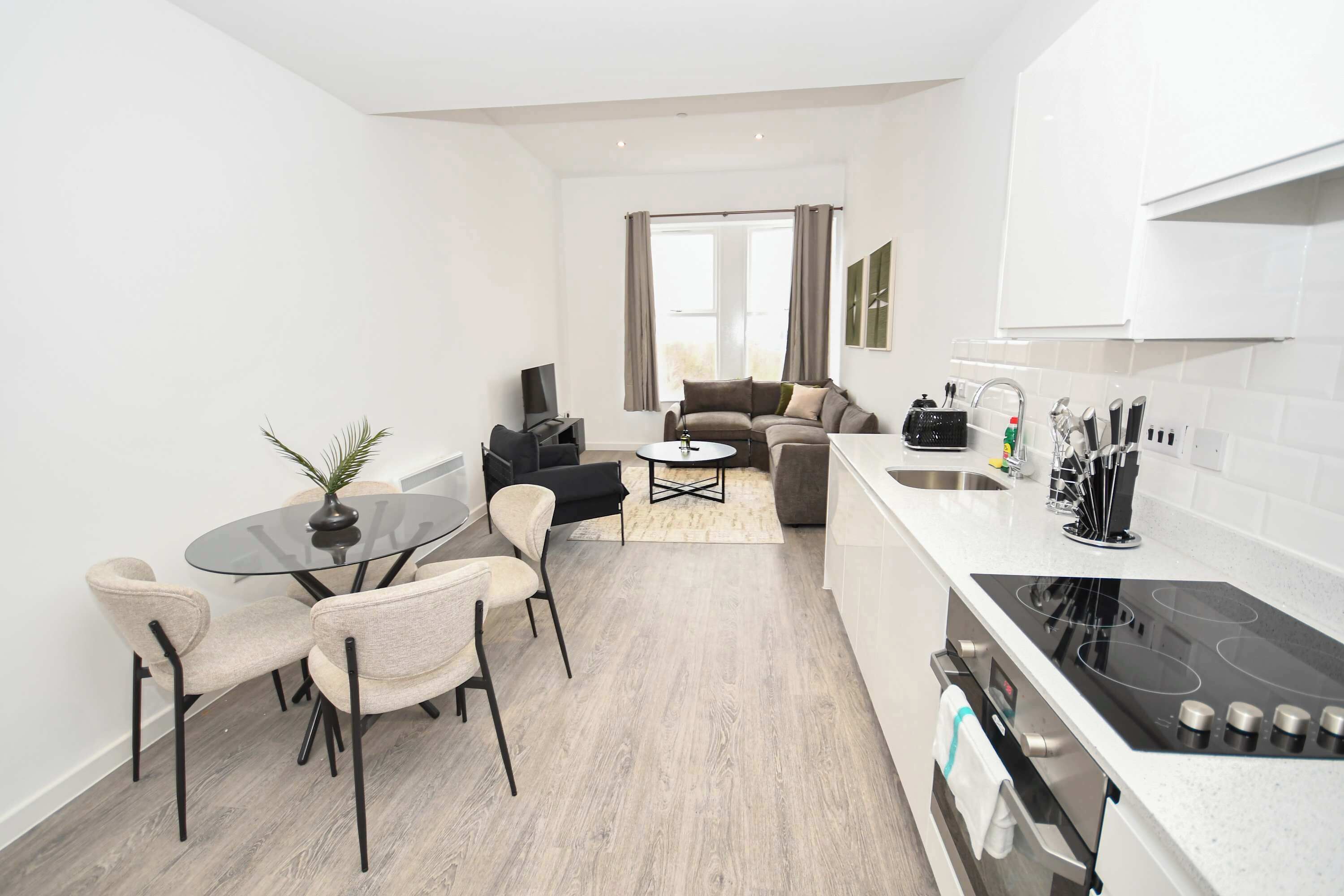 Cozinha moderna com fogão preto, área de refeições com mesa e cadeiras de vidro, e sala de estar com sofá e televisão.