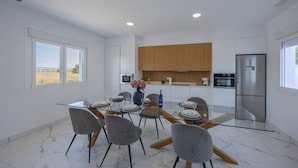 A modern kitchen with white cabinets, a stainless steel refrigerator, and a dining area with a glass table and grey chairs. - Psicomagia Deluxe Villa by Fidalsa (Dolores)
