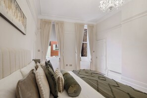 A bedroom with a large bed, beige curtains, a chandelier, and a view of a brick building. - Upscale Central London Apartment (London)