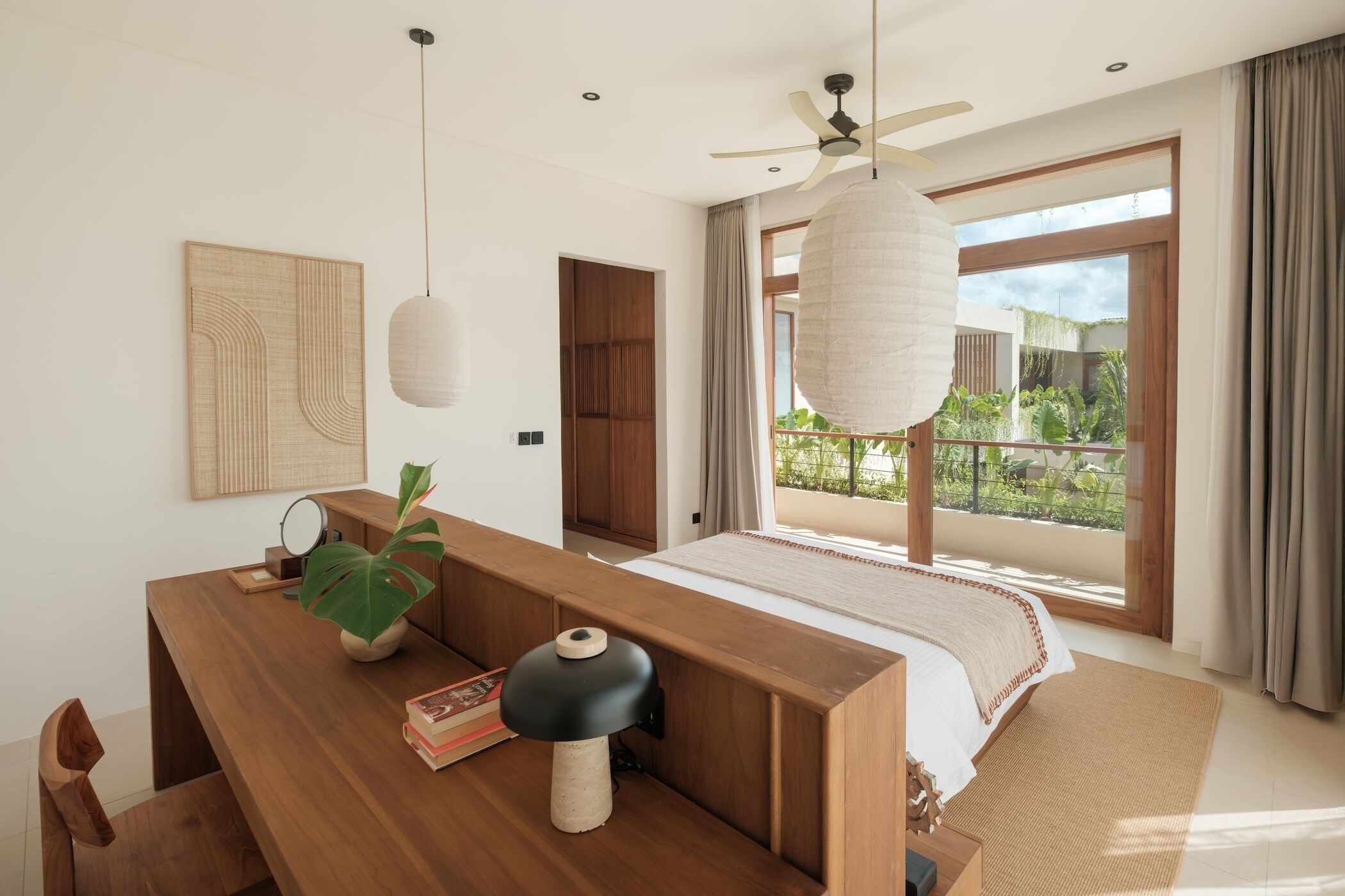Un dormitorio moderno con un escritorio de madera, un ventanal con vistas a la vegetación, un ventilador de techo y una cama con cubrecama blanco.