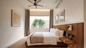 A modern hotel room with a large bed, wooden headboard, bedside table, and a view of palm trees through the window. - Manu Villas (Denpasar)