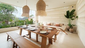 A modern dining area with a wooden table set for four, two hanging pendant lights, a white cushioned bench, and a view of greenery outside. - Manu Villas (Denpasar)