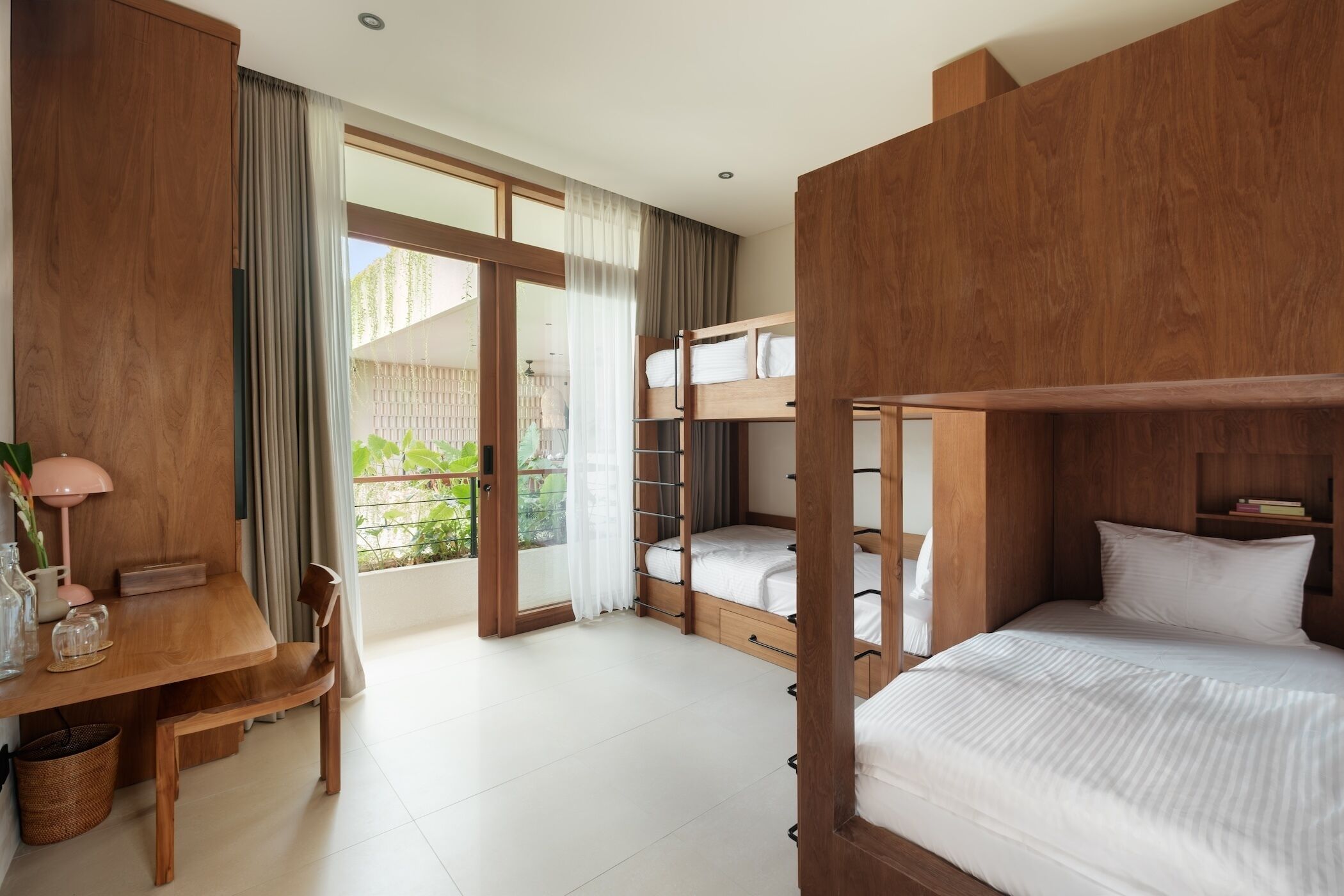 A room with bunk beds, a wooden desk with a chair, a small table with a lamp, and a view of greenery through the door.