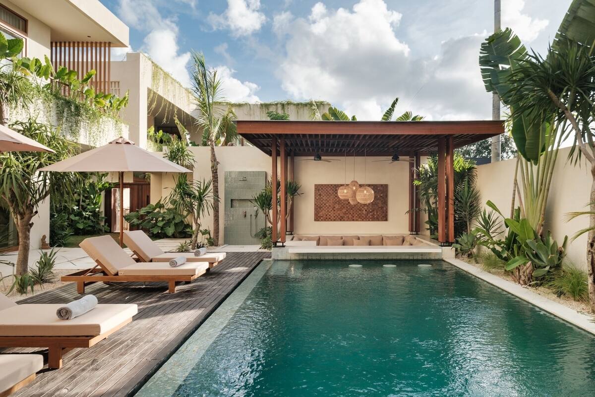 A swimming pool with lounge chairs and umbrellas, surrounded by tropical plants and a covered seating area.
