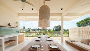 A dining area with a long table set for six, a hanging light fixture, and a view of greenery outside. - Manu Villas (Denpasar)