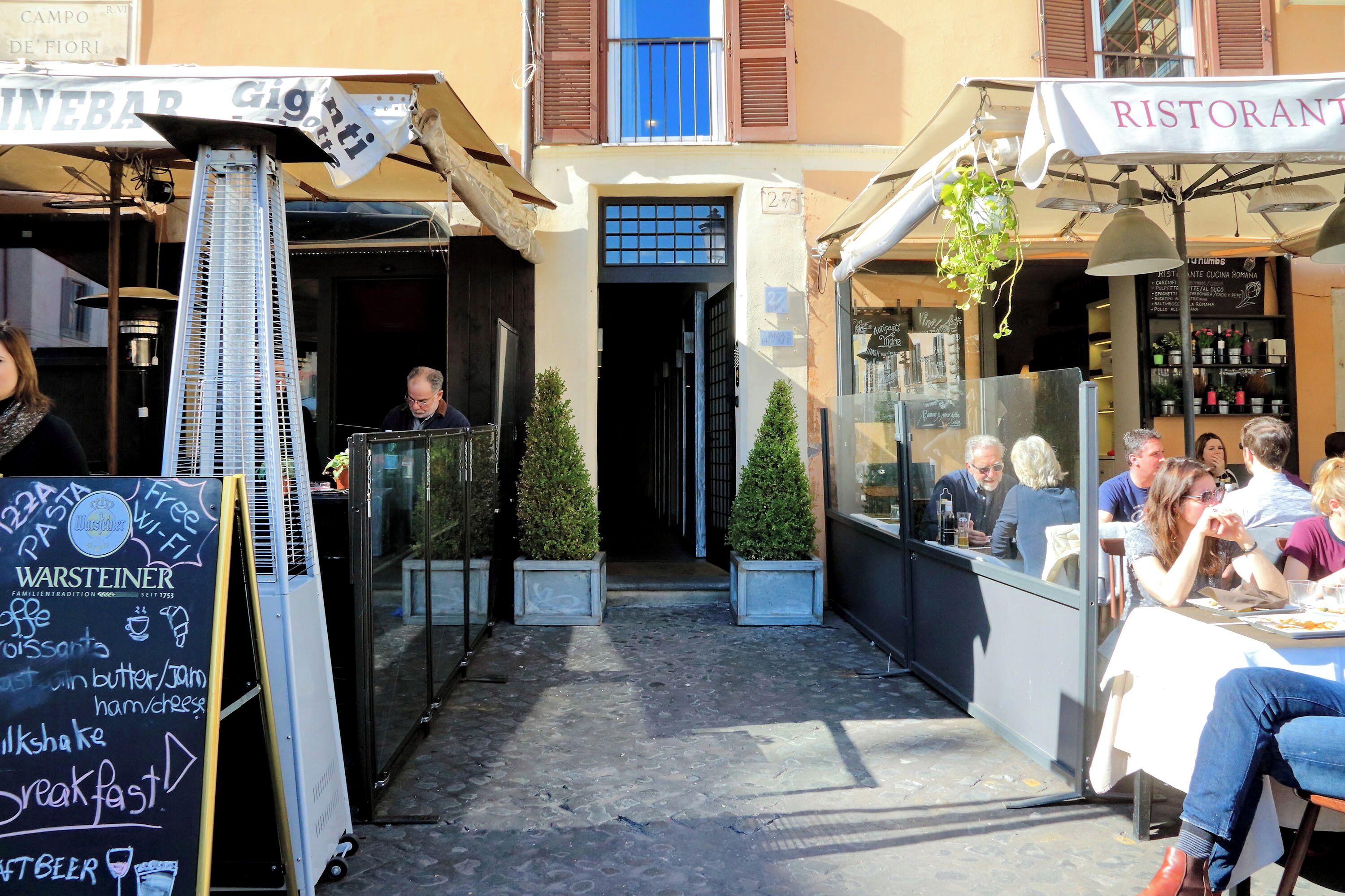 Un área de café al aire libre con mesas y sillas, un menú en pizarra y un cartel que anuncia un restaurante cercano.