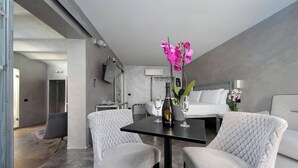 Una habitación de hotel moderna con una cama, dos sillones blancos tapizados, una mesita con una botella y vasos, y un jarrón con flores rosadas.