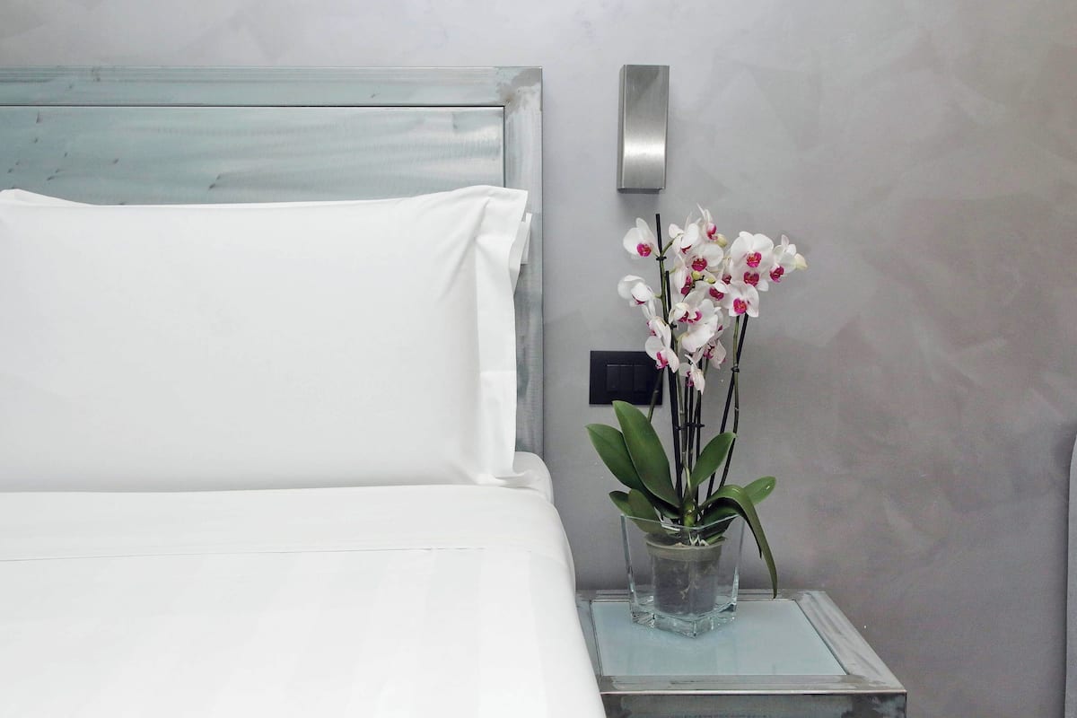 Una cama bien hecha con ropa blanca, una mesita de noche con un florero de vidrio transparente que contiene orquídeas blancas, y una lámpara moderna fijada a la pared.