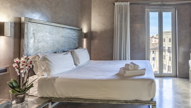 Habitación de hotel con una cama grande, una mesita de noche con un florero y vistas a edificios a través de una ventana, además de un balcón.