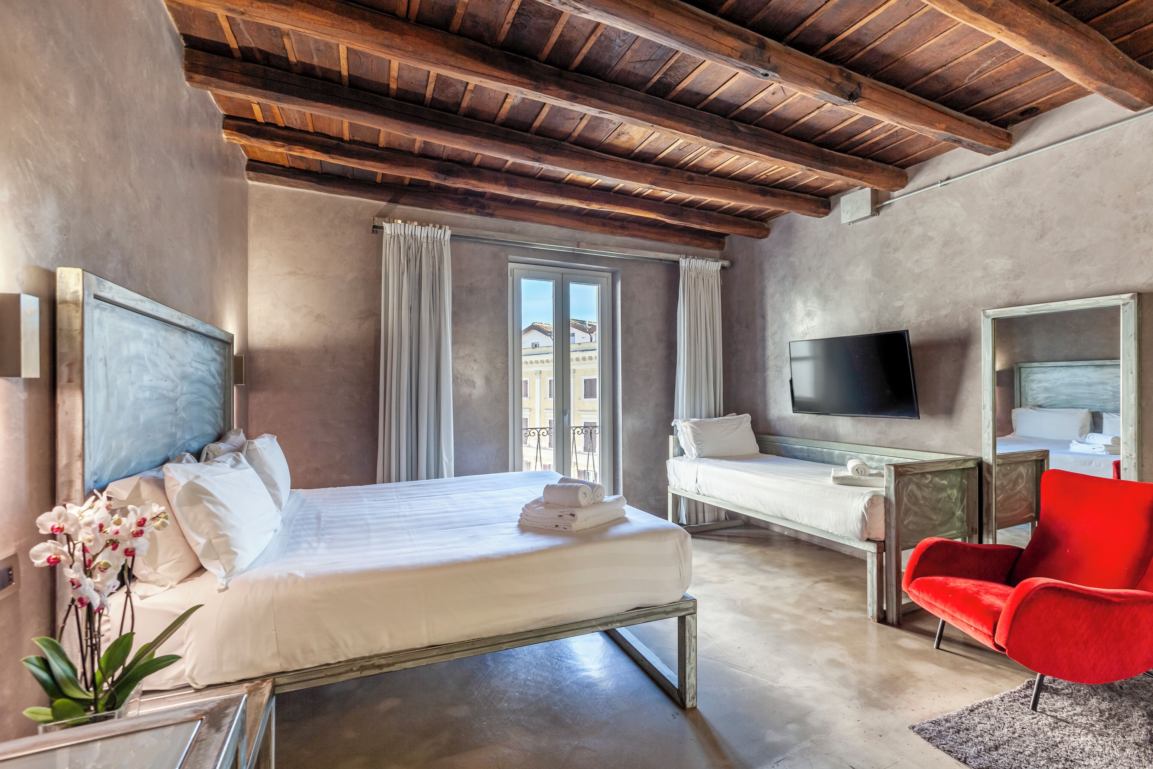 Habitación de hotel con cama, escritorio, silla roja, televisión y ventana con cortinas.