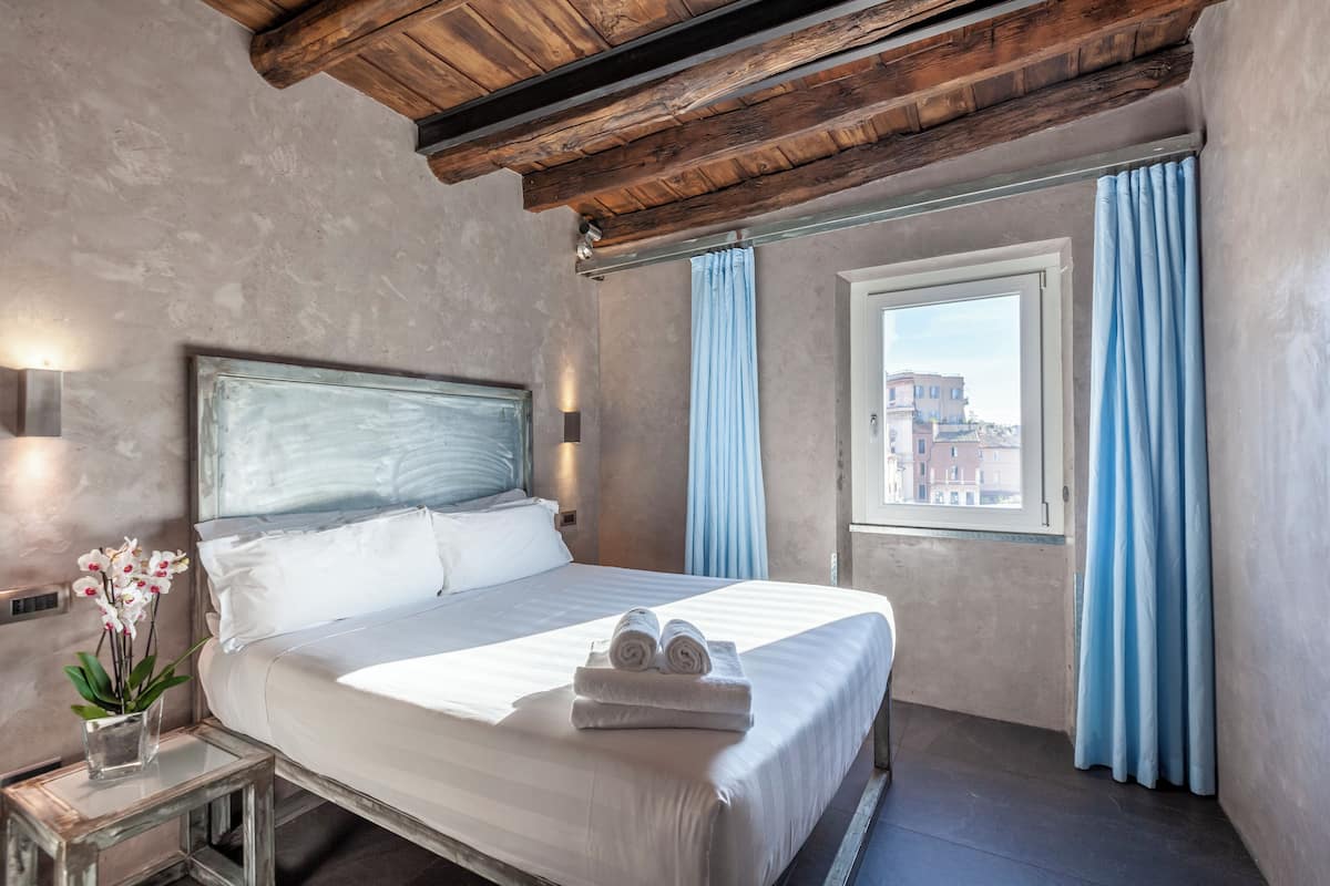 Un dormitorio con una cama grande, techo de madera, ventana con cortinas y vistas a edificios.