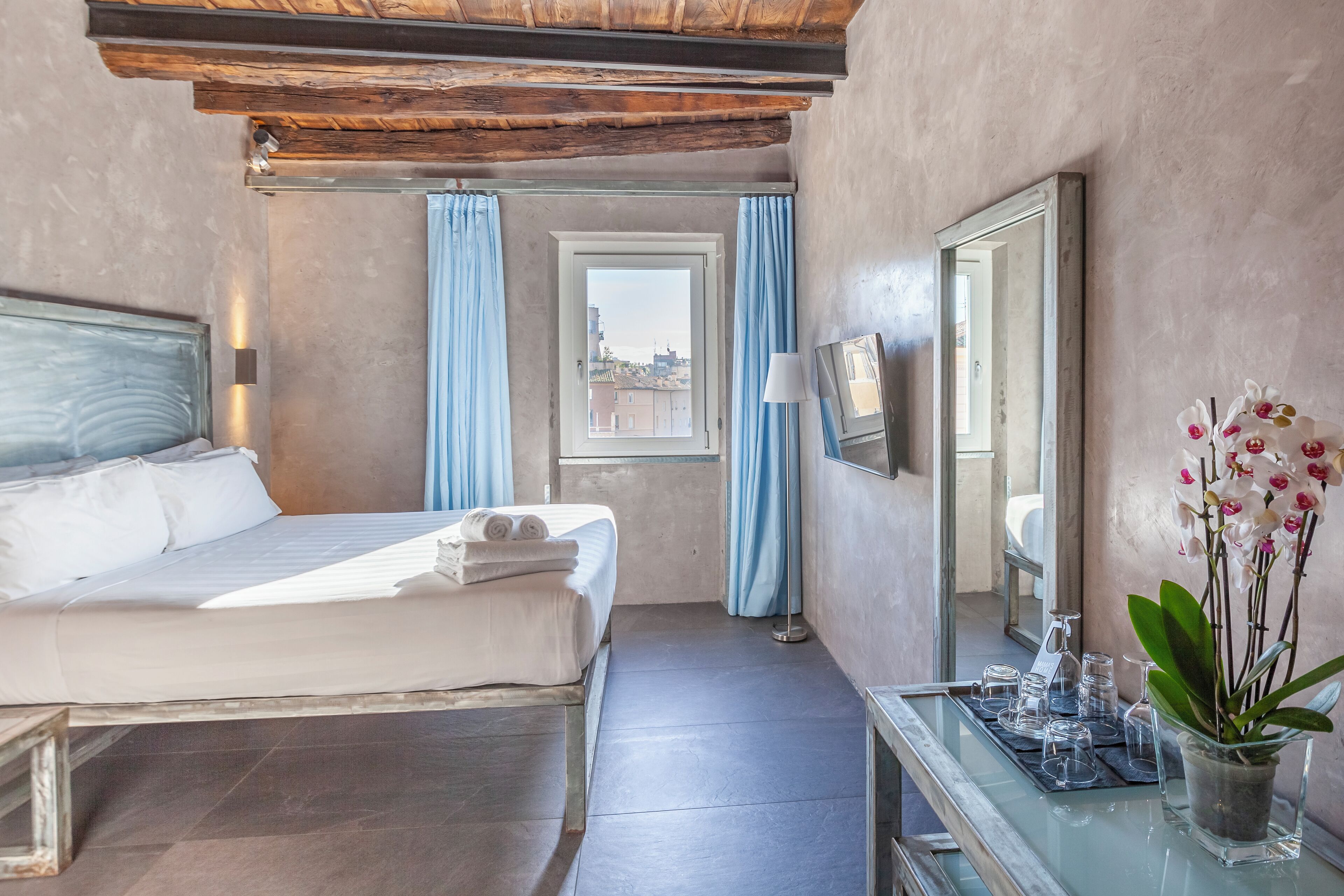 Habitación de hotel con una cama, una mesita de noche con lámpara, un espejo y una ventana con cortinas.