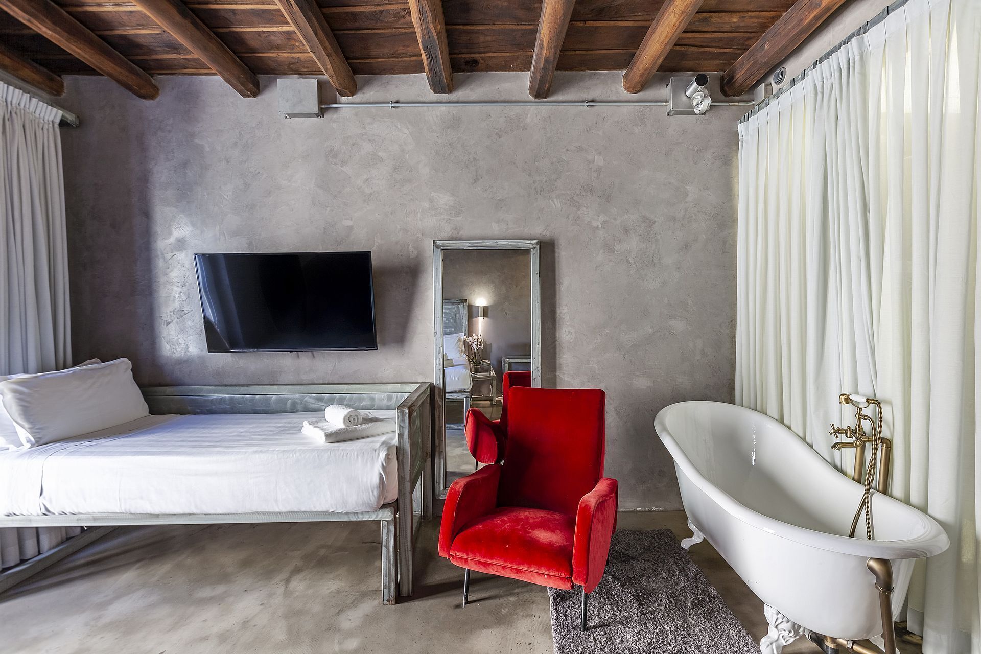 Une chambre d’hôtel moderne avec un lit, un fauteuil rouge, une baignoire blanche et un téléviseur à écran plat fixé au mur.