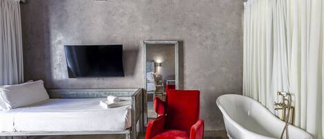 Une chambre d’hôtel moderne avec un lit, un fauteuil rouge, une baignoire blanche et un téléviseur à écran plat fixé au mur.