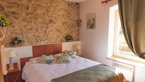 Une chambre avec un mur en pierre, un lit avec de nombreux oreillers, une table de chevet avec une lampe et une fenêtre avec des rideaux.