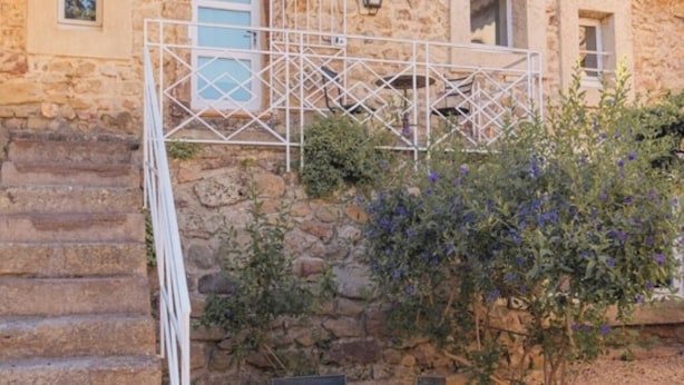 Un bâtiment en pierre doté d’un balcon en métal blanc, entouré de plantes et d’une zone recouverte de gravier.