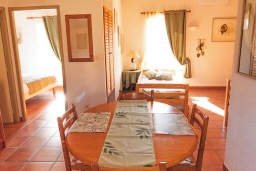 A dining area with a round table and chairs, a bed with a green curtain, and a window with a view.