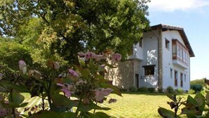 A white building with a stone base and a tiled roof, surrounded by greenery and flowering plants. - la casona encanto rural (Llanes)