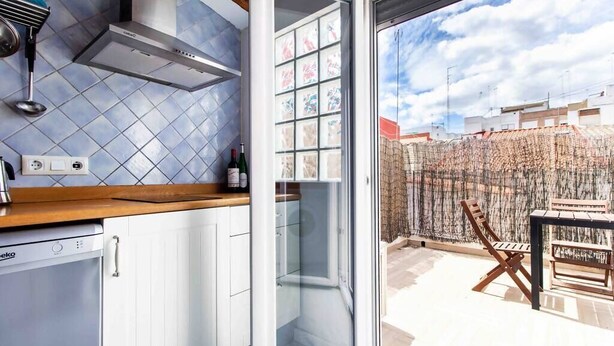 A kitchen with a dishwasher, a stove hood, and a window overlooking a courtyard with a chair and table. - Studio for couples with terrace on the beach (València)